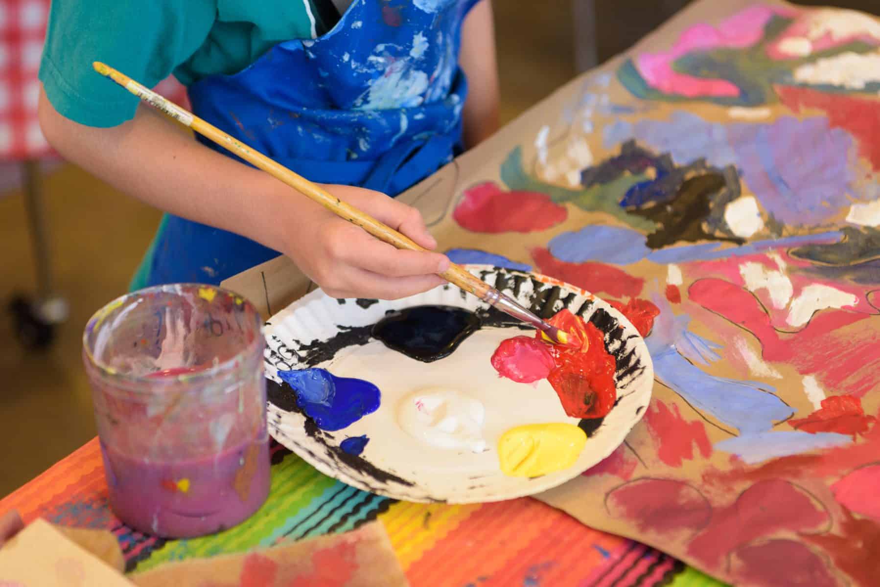 A close-up view of a child's hand holding a paint brush above a paper plate filled with paints of different colors.