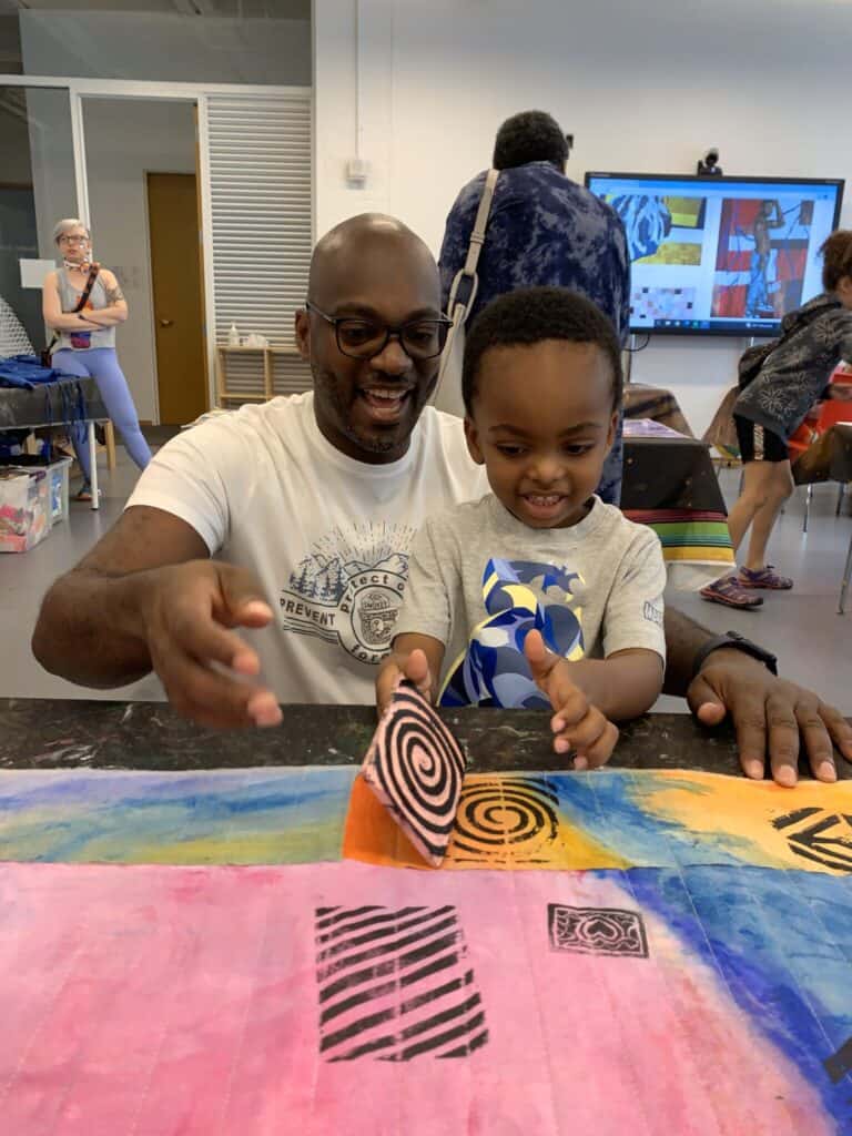 A person and a child use a relief print to stamp patterns on a colorfully painted surface. 