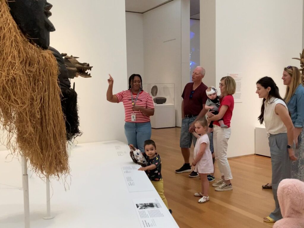 A group of adults and children in an art gallery gather around a person gesturing to three African masks on display. 