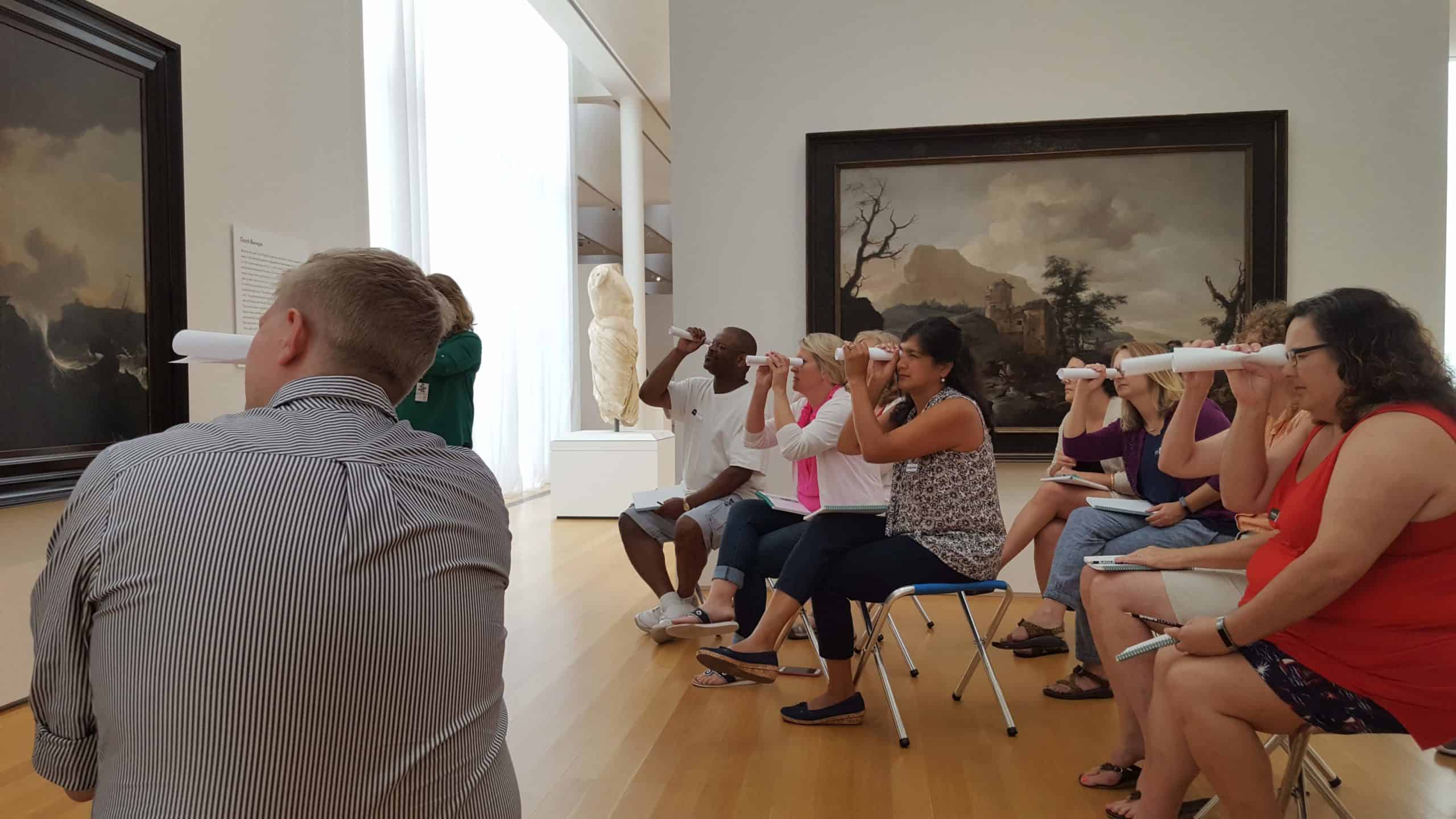 People sit on stools in a gallery and view a painted artwork through rolled up pieces of printer paper. 