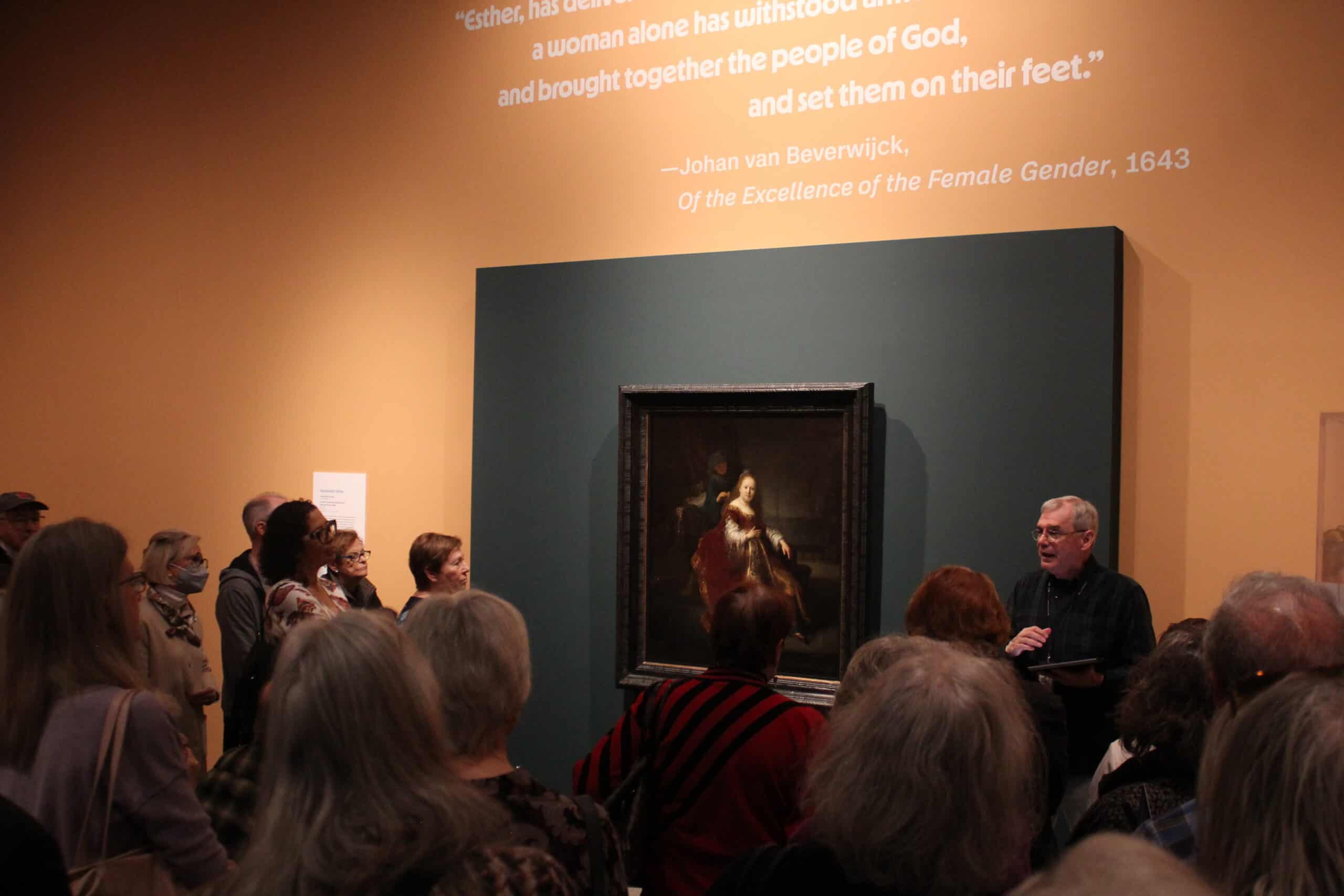 A man speaks to a large group of people gathered around a painted portrait. 