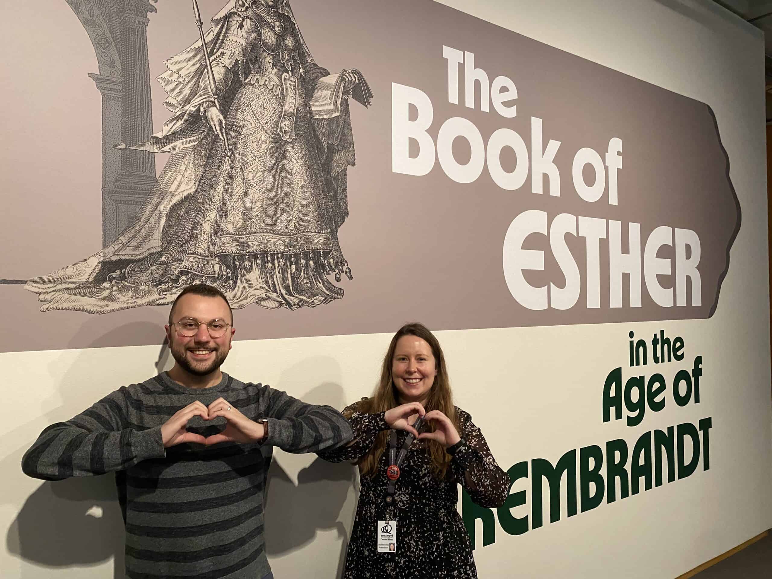 Two people making the shape of a heart with their hands stand in front of a beige and gray wall with the words "The Book of Esther in the Age of Rembrandt" printed on it. 