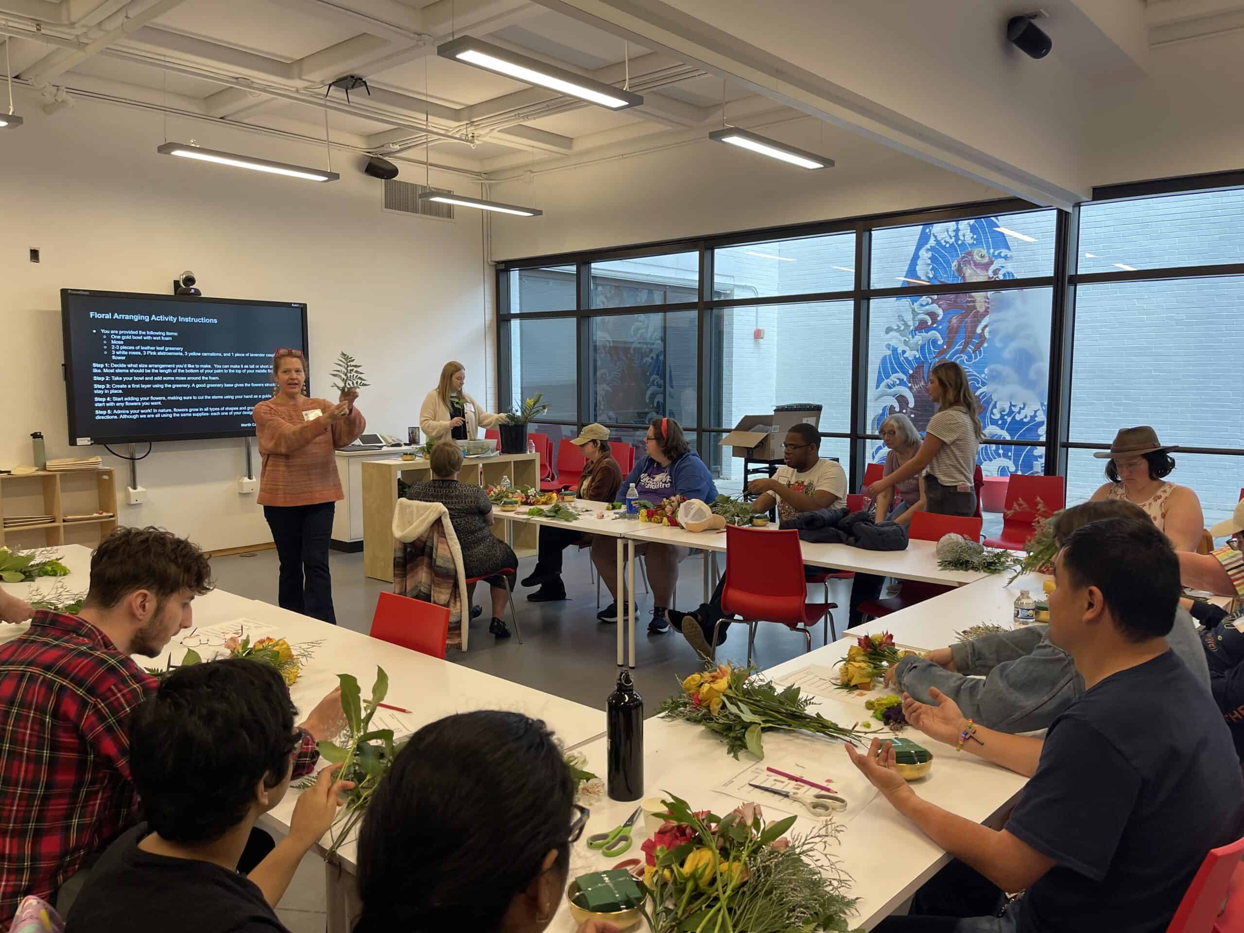 Two floral designers stand in front of workshop participants who are making floral arrangements.
