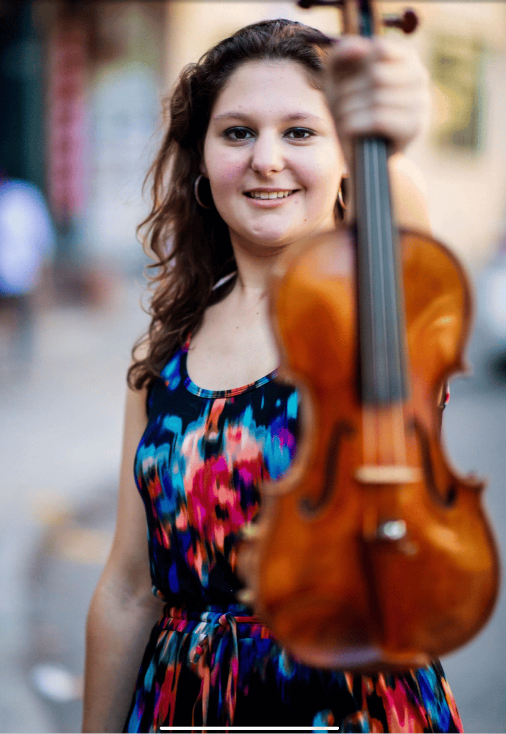 A smiling woman in a colorful sundress holds her violin by the neck and shows it to the viewer.