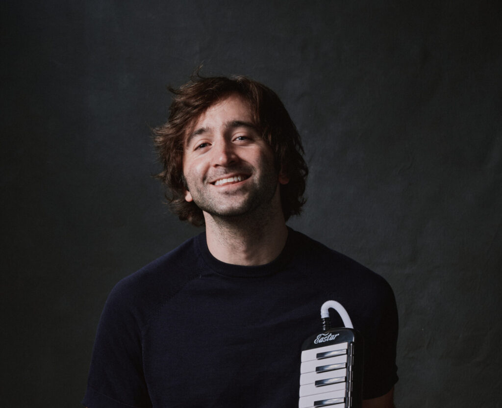 A brown-haired smiling man wearing gray pants and a black T-shirt holds a small piano keyboard.