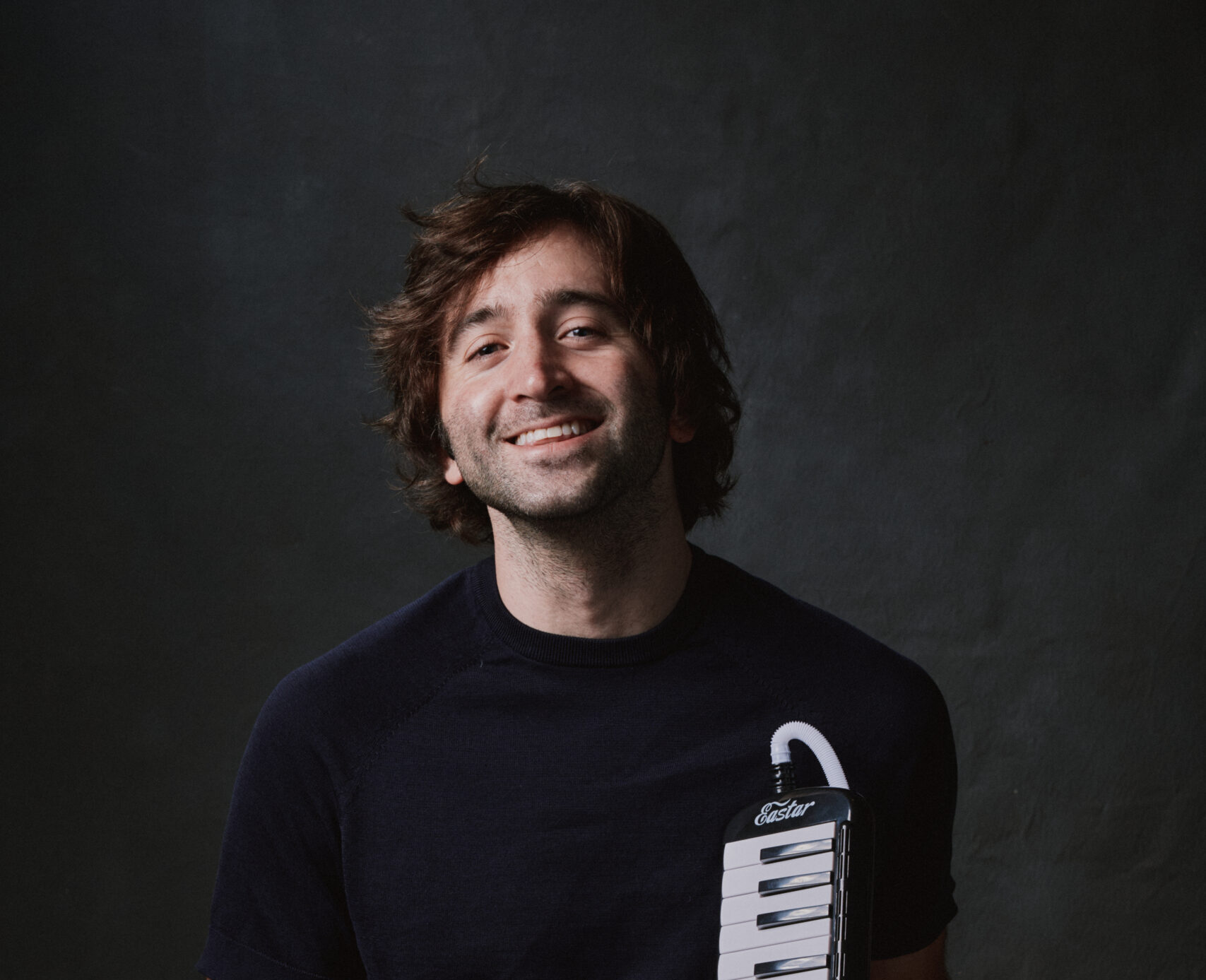 A brown-haired smiling man wearing gray pants and a black T-shirt holds a small piano keyboard.