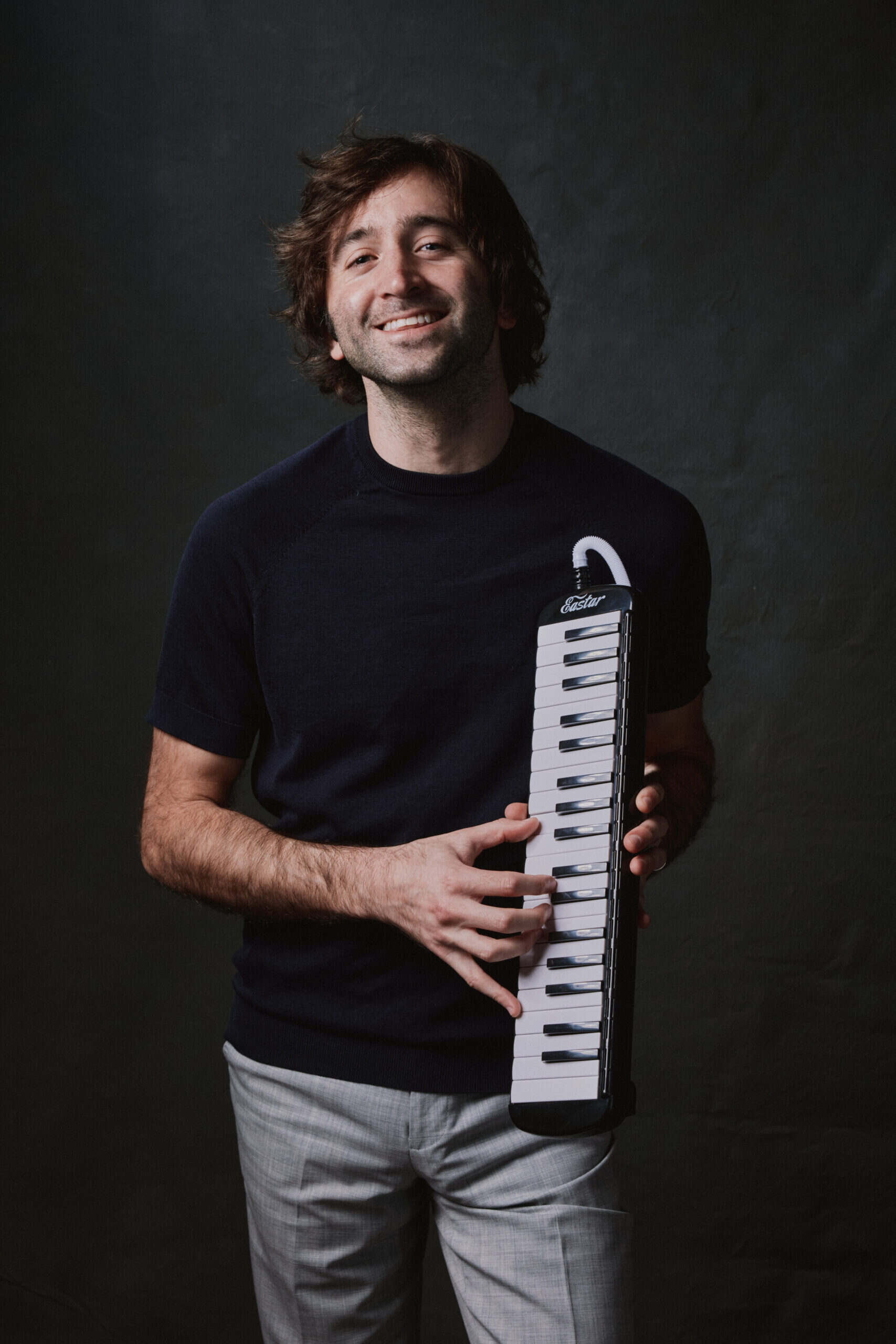 A brown-haired smiling man wearing gray pants and a black T-shirt holds a small piano keyboard.