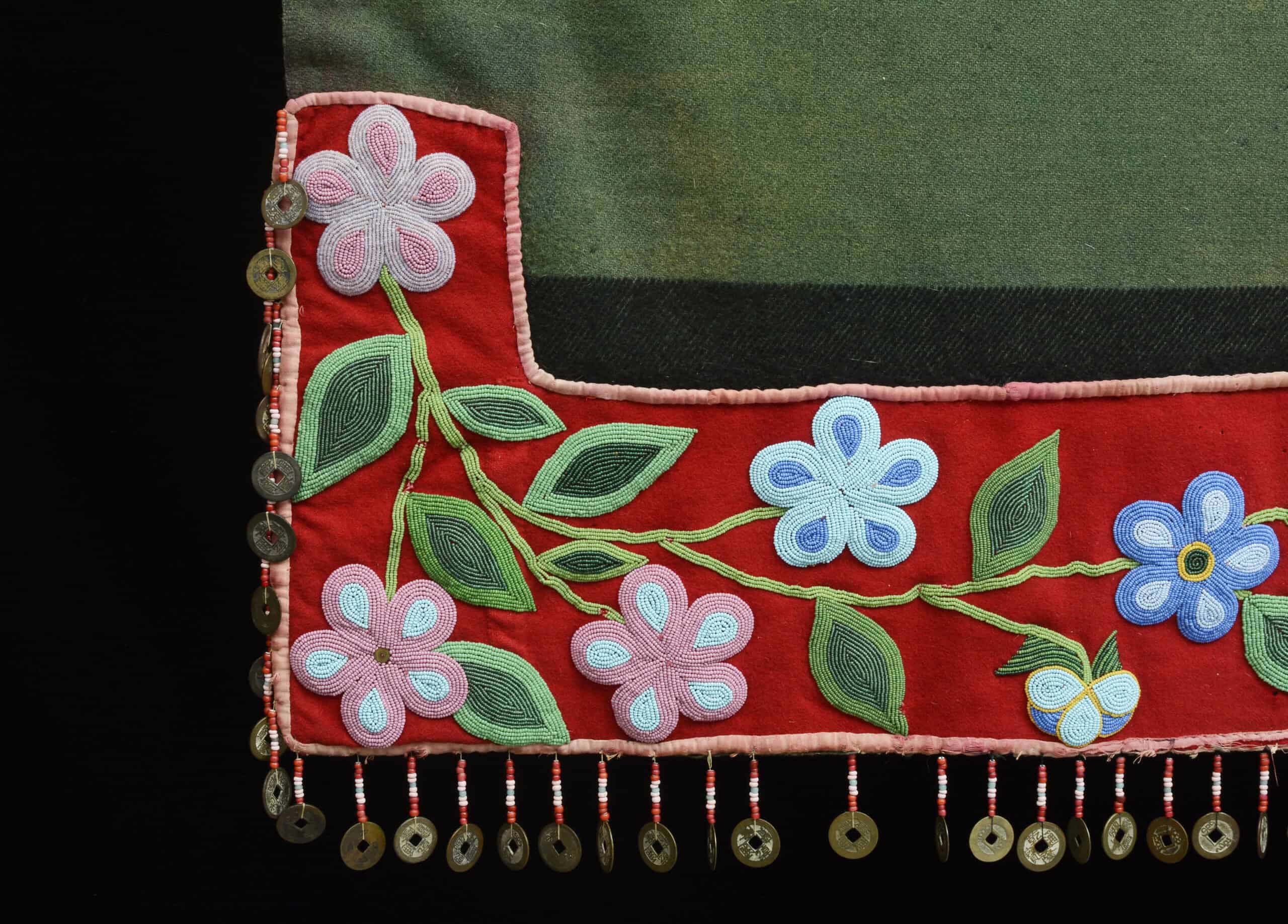 The corner of a green blanket with red trim embroidered with beads and a fringe edge with coins.