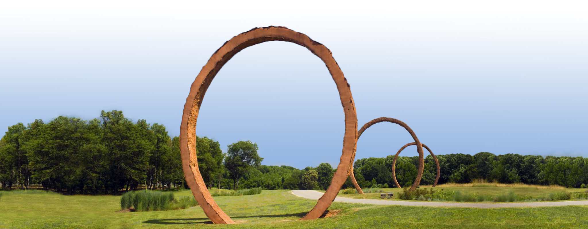 Three large, red-orange, ring-shaped sculptures on a grassy lawn. There is a paved path between two of the rings. In the background there are trees and a blue sky.