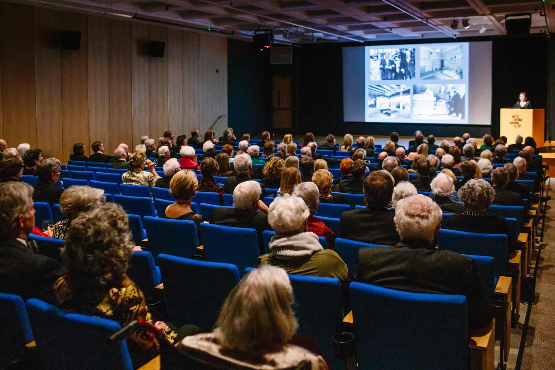 A group of people sitting in an auditorium and watching a person give a presentation from behind a podium. Four indistinct black-and-white images are projected on the auditorium screen.
