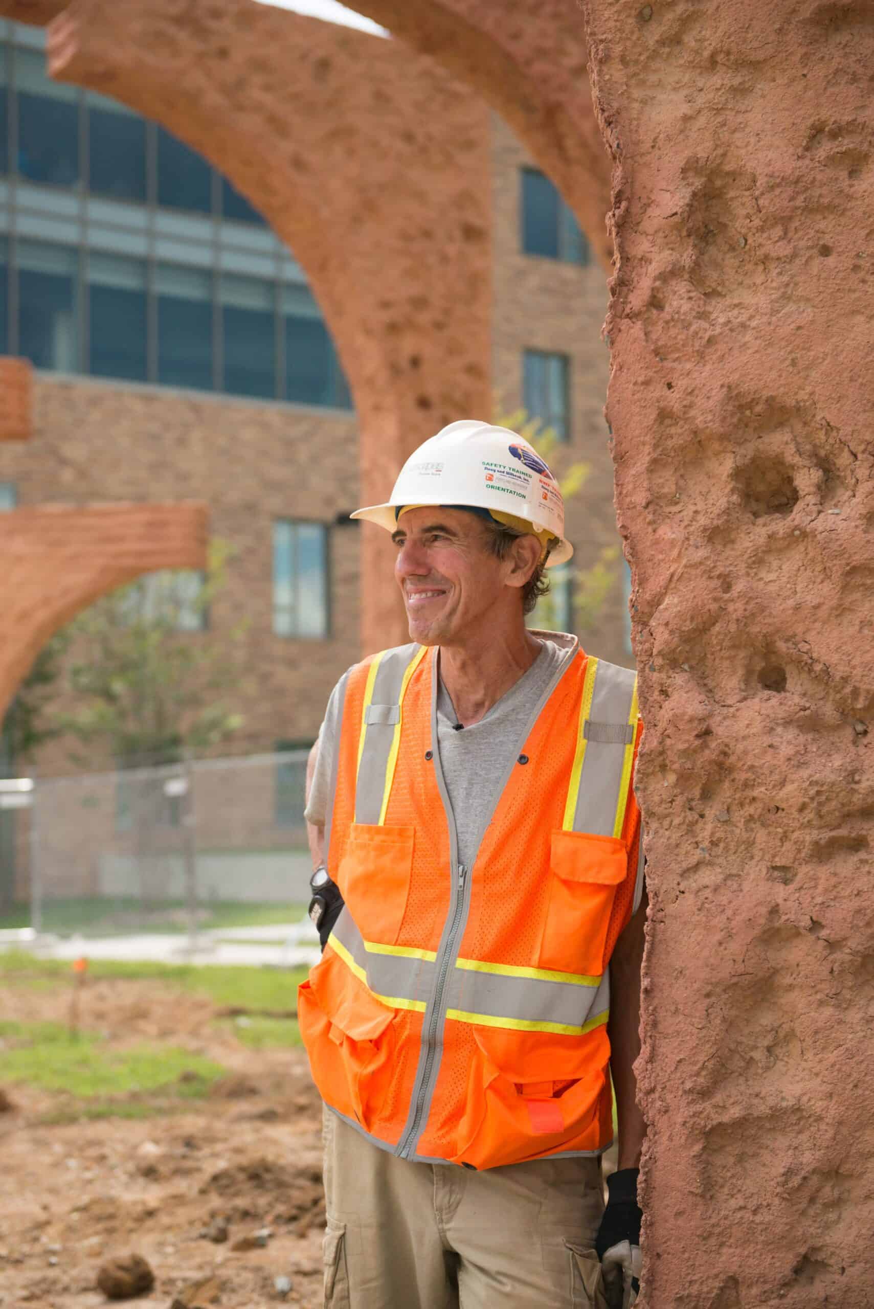 A white, tanned man in a white construction hat and neon orange construction crew vest leans against a terracotta ring of an earth-cast sculpture.