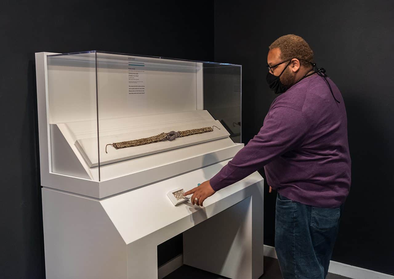 A gentleman reaches out to touch a box of shells built into a white podium that houses a work of art. He is looking at the work of art. 