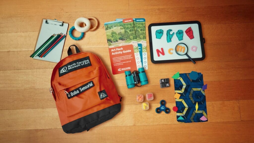 Various games, activities, and informational resources spread out on a table next to an orange backpack with the words "North Carolina Museum of Art" printed on it. 