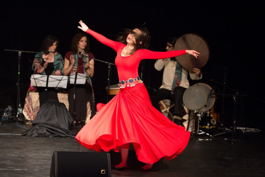 A woman in a long red dress twirls around with her arms out while two woman clap and a percussionist plays in the background.