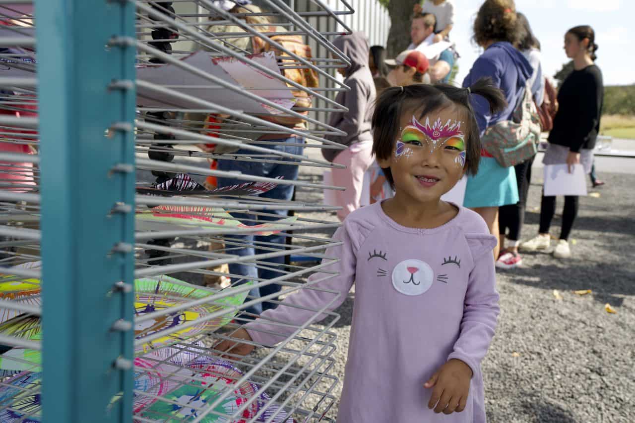 Child wearing face paint poses in front of an outdoor art drying rack.