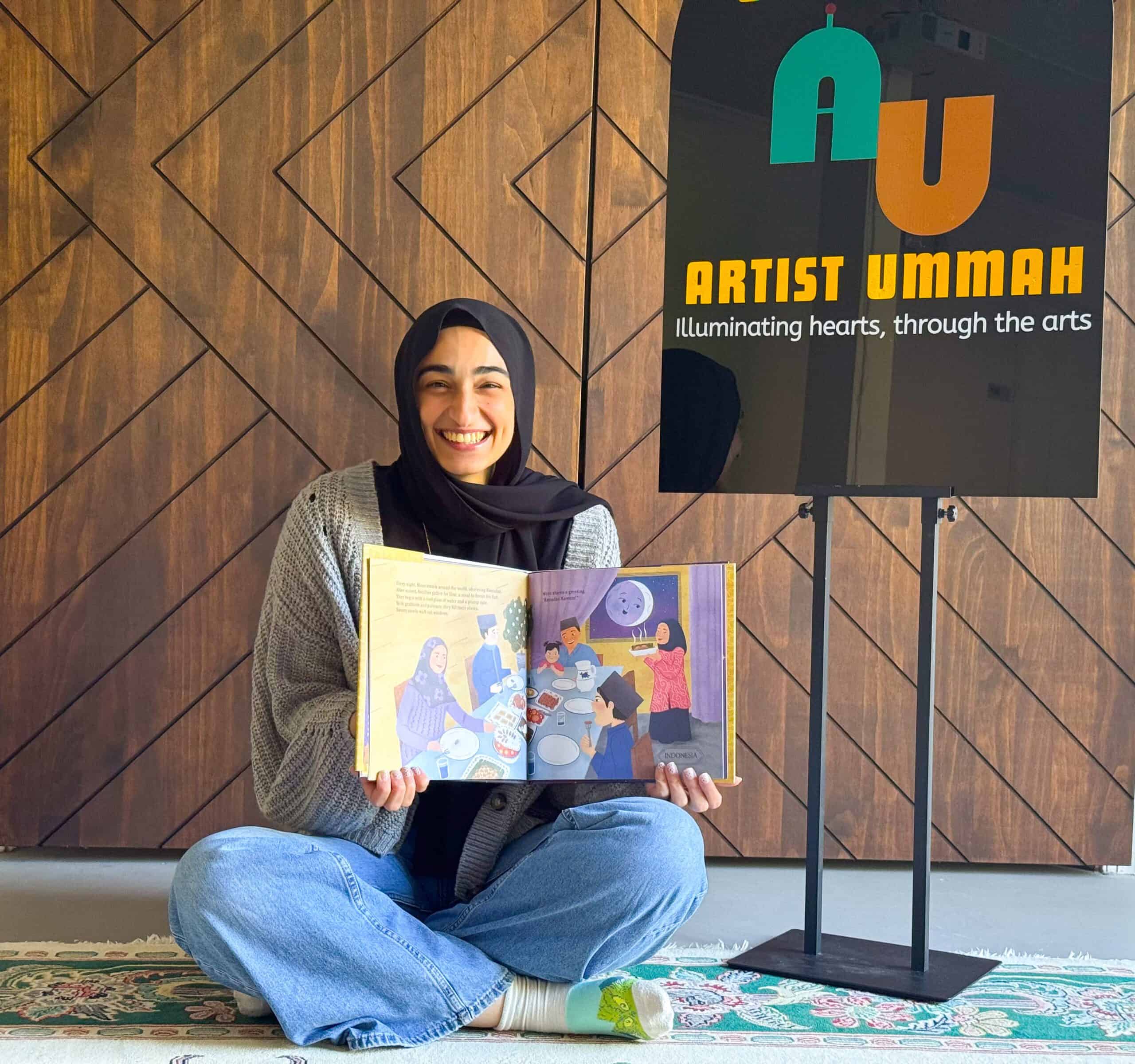 A smiling woman in a hijab sits cross-legged on the floor and holds open an illustrated book beside an Artist Ummah sign.