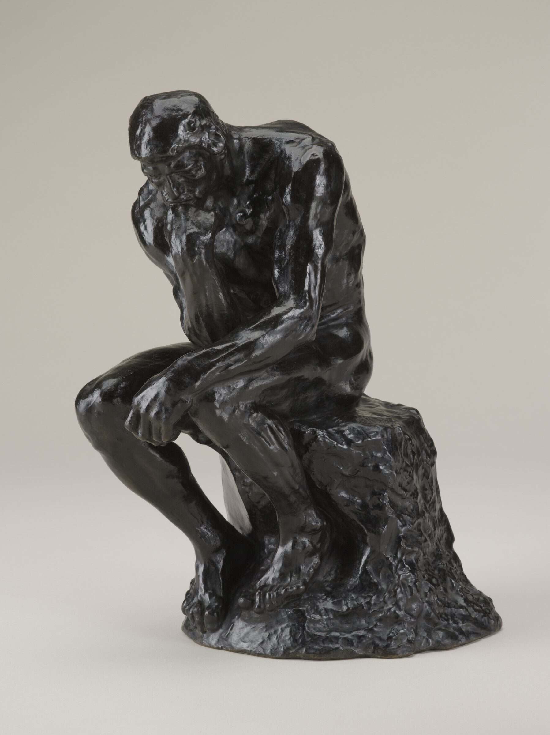 Bronze sculpture of a nude man sitting on a rock. His chin rests on his right hand, and his right elbow rests on his left thigh.