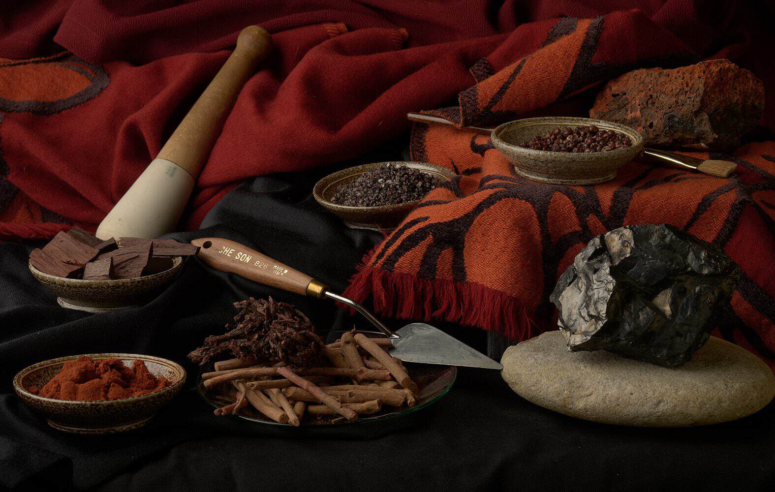 A photographic still life that includes red drapery, pestle, spatula, several large rocks, and bowls filled with organic materials.