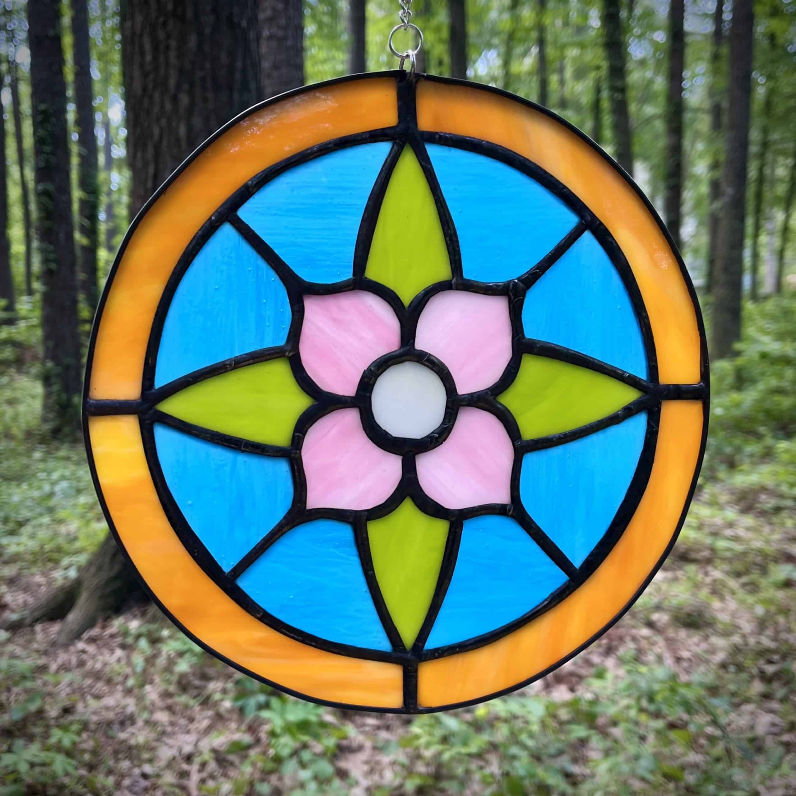 A circular stained glass suncatcher featuring a floral design in a pink, blue, green, and yellow color palette.