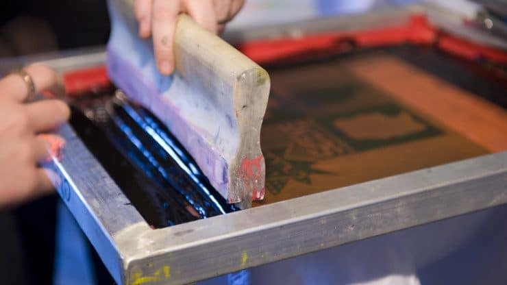 One hand holds a silkscreen while another hand holds a squeegee perpendicular to it. Black ink pools on one side of the squeegee.
