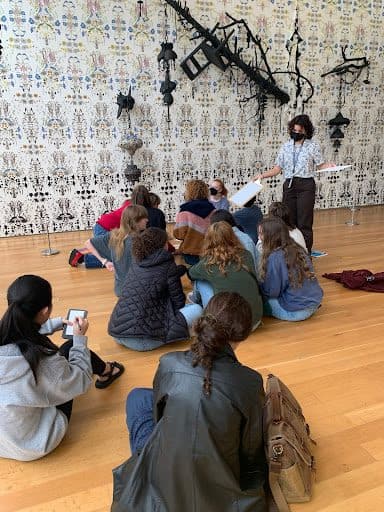 A group of teens sit on a wood floor in front of a wall art installation. A person wearing a face covering holds a clip board out to the group.