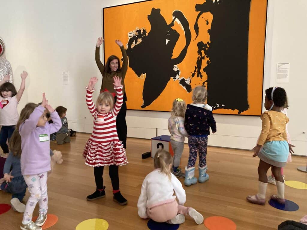 A group of children play in front of an abstract painting of two bold black forms floating on a warm orange field, with ragged edges and white flecks suggesting motion and contrast. A person with their arms in the air stands in front of the children. 