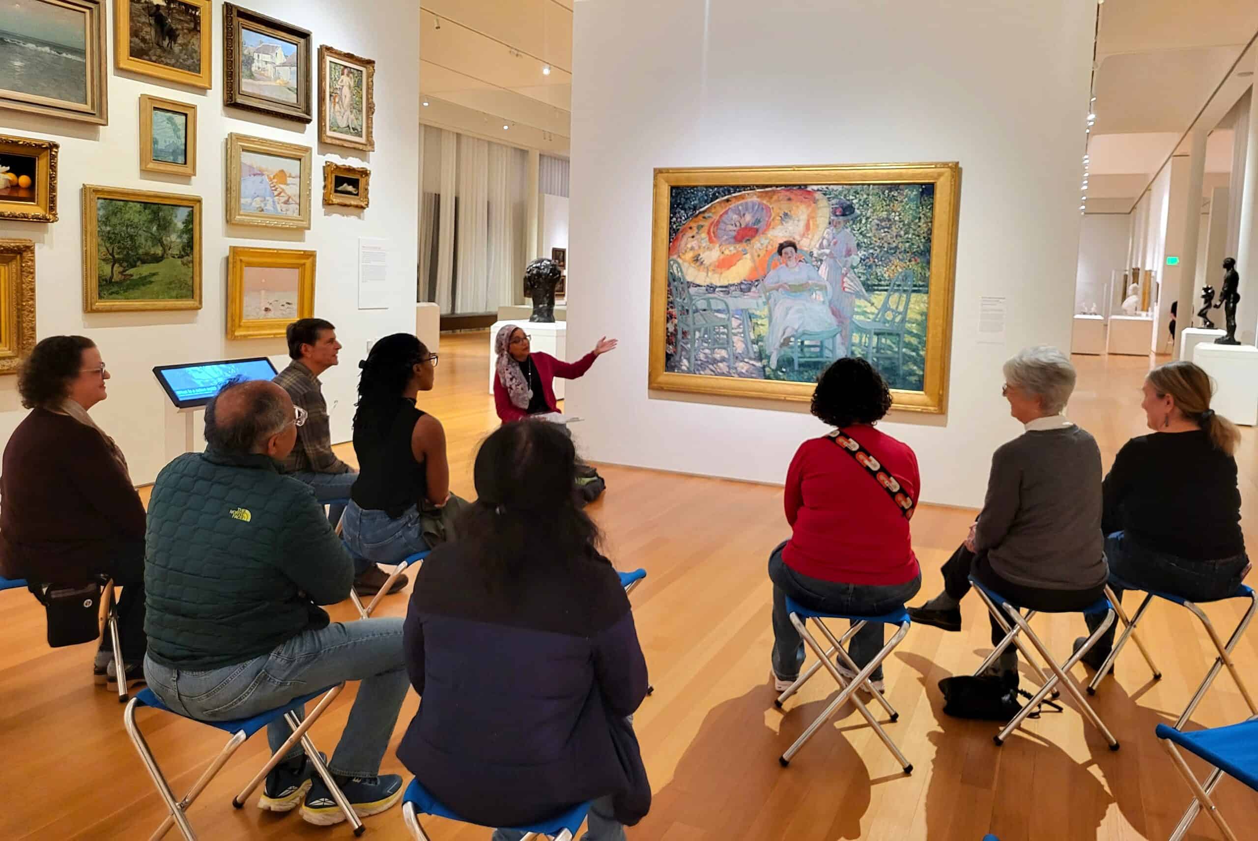 A group of people seated on stools in a gallery look at a large framed painting of a woman under a large parasol.