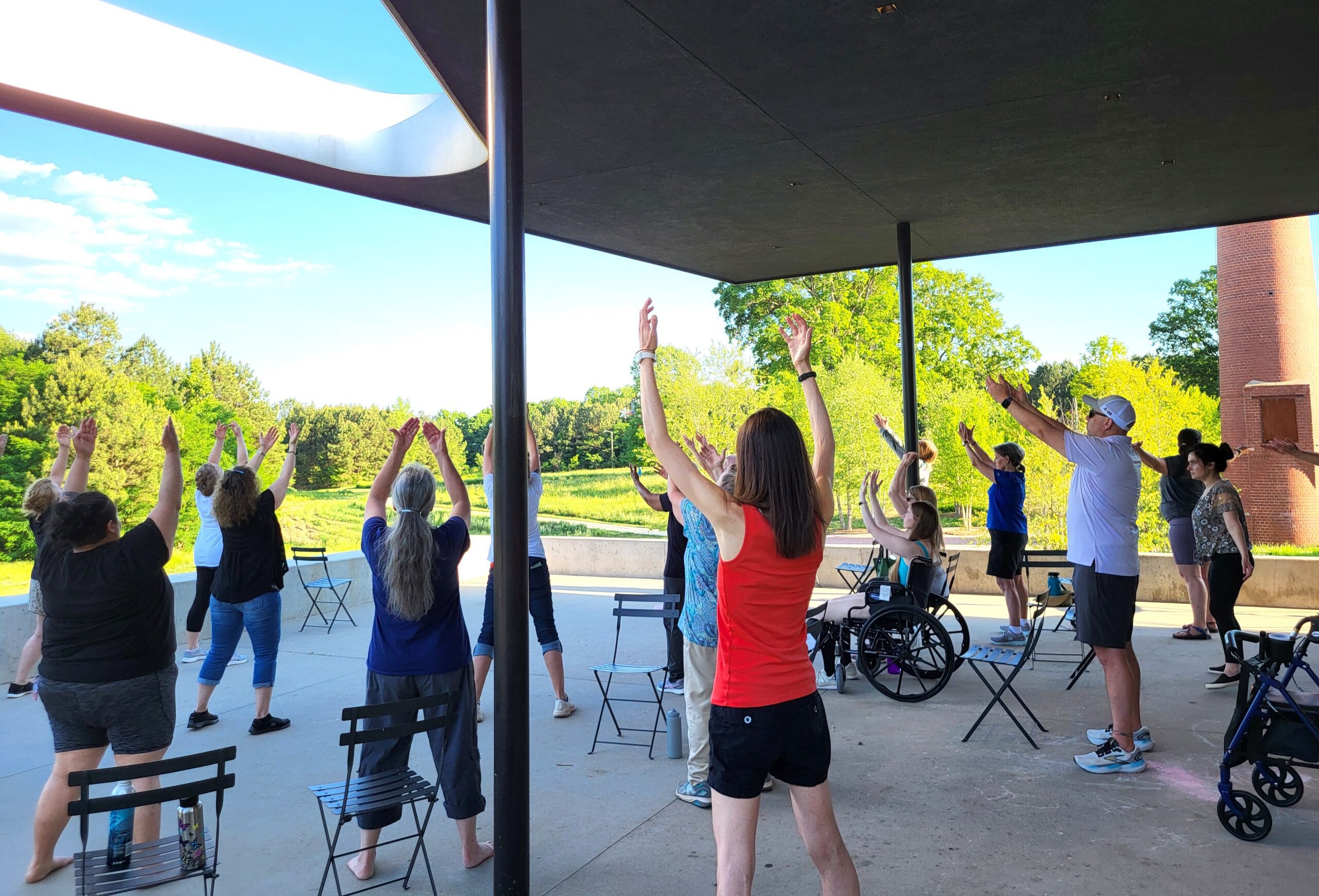 A group of people stand and sit on an outdoor patio with their arms raised over their heads and face a bright green tree line.