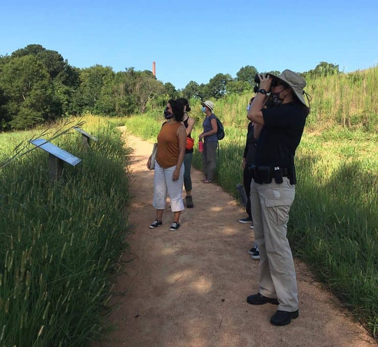 A group of six people wearing face coverings stand on a dirt path in the middle of a tall grass meadow and gaze out of frame. One person holds binoculars to their eyes. 
