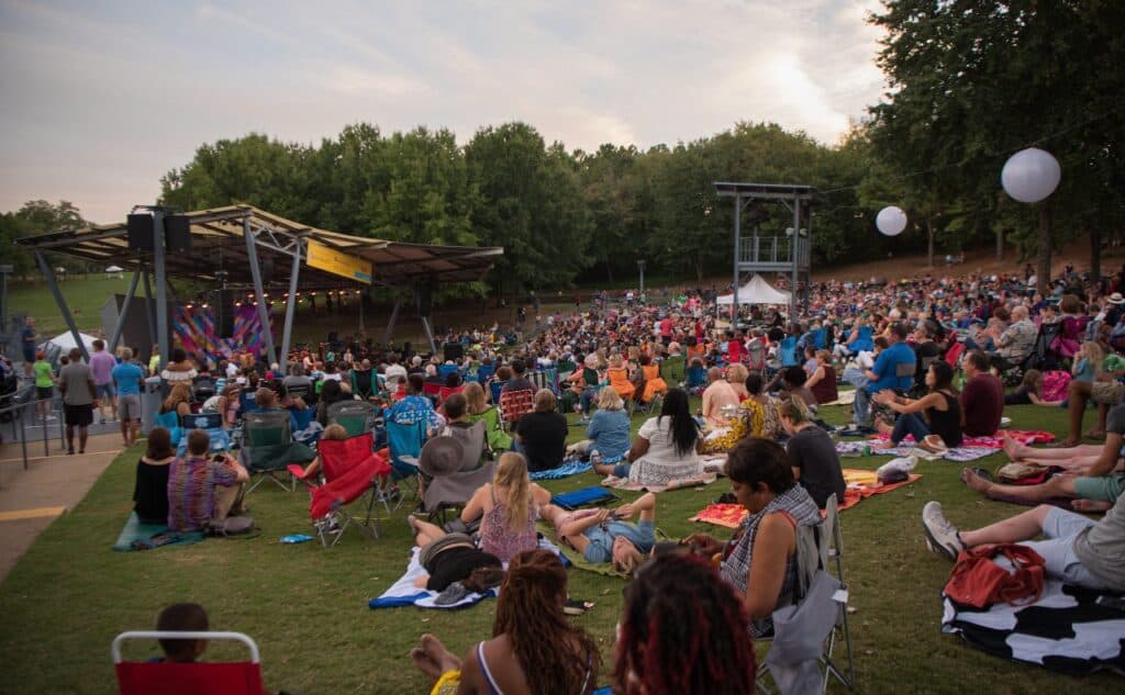 A crowd of people seated on a sprawling outdoor lawn face a covered amphitheater stage. In the background there are trees set against a dusky sky.