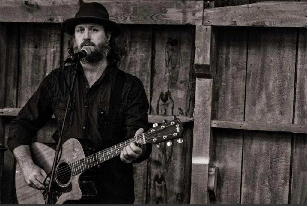 A man in a hat holding a guitar stands before a microphone and against a tall wooden fence.