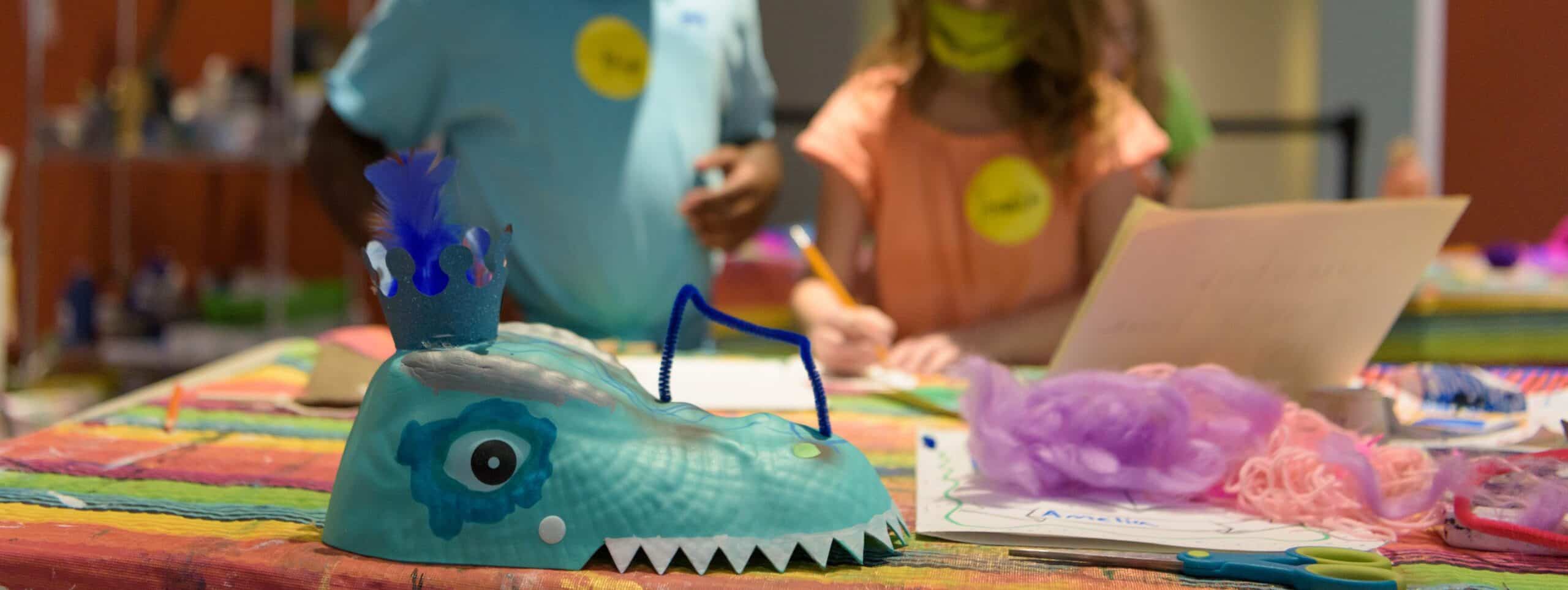 A mask-like sculpture sits on a table in front of two young children making art.