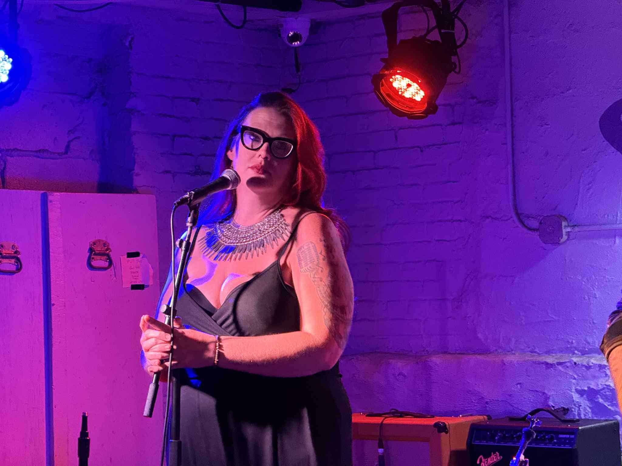 A singer wearing a black dress stands in front of a microphone and is lit by purple and blue lights.