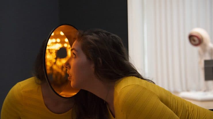 A person bends over to look inside a large mirror-covered box filled with yellow light. 