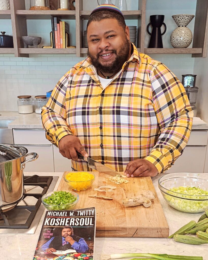 A man wearing a yarmulke and plaid shirt stands behind a kitchen counter and looks up from chopping vegetables.