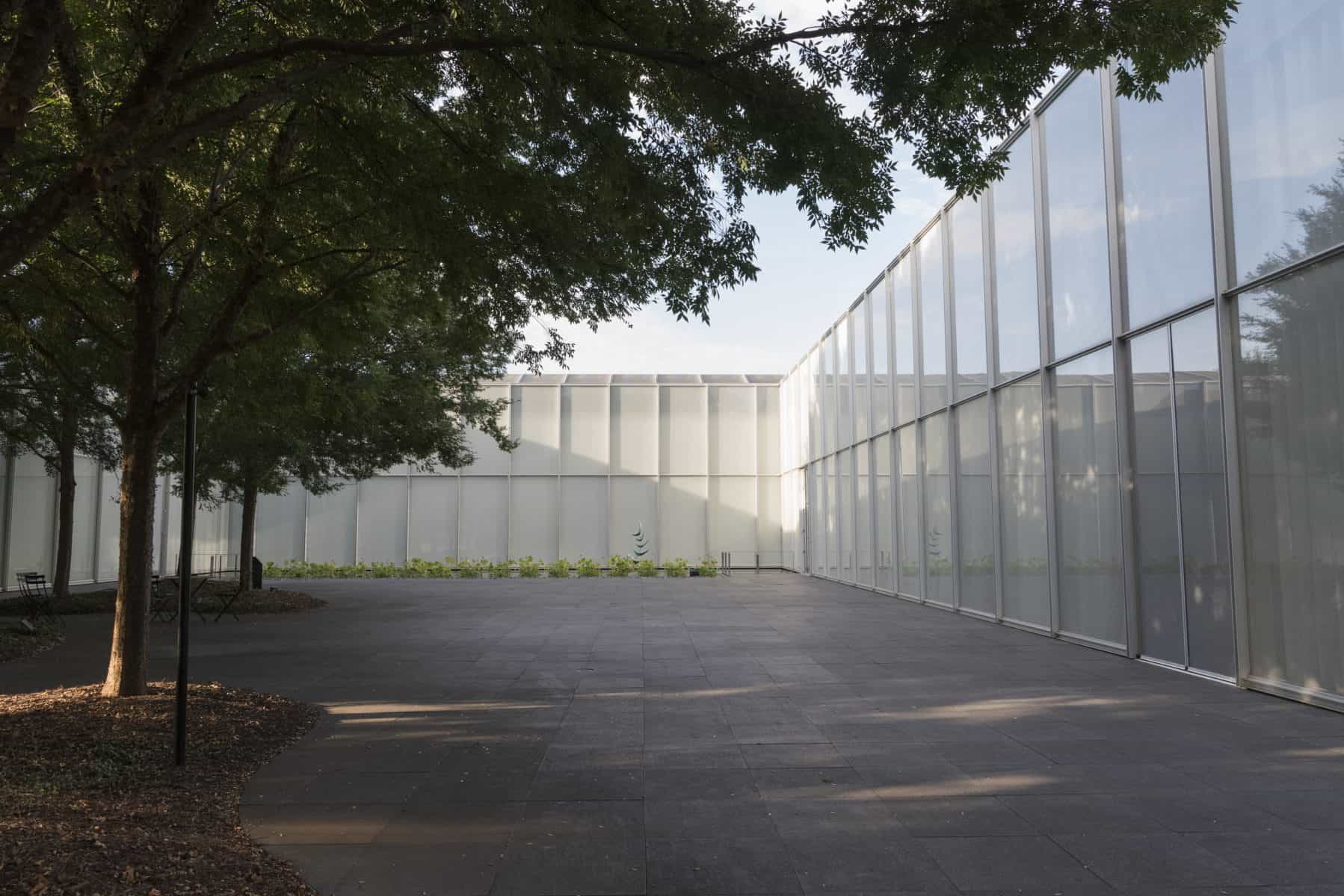 A courtyard outside a modernist, glass and aluminum building. In the foreground there are trees providing shade to the courtyard, and in the background there is a reflecting pool and an abstract sculpture.
