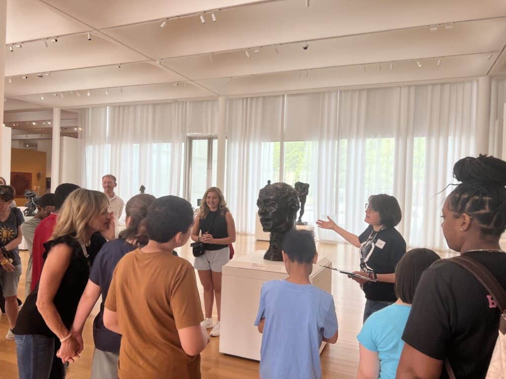 Two women flank a bronze sculpture of a head, with a group of people standing in front of them.