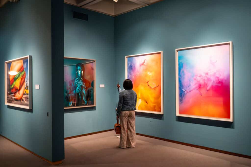 Two people in an indoor art gallery looking at colorful large photographic prints on blue walls.