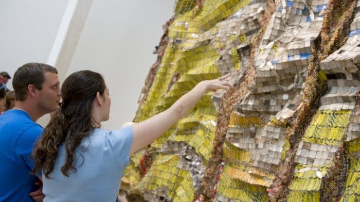 Two people stand in front of and gesture toward a giant, clothlike metal sculpture with yellow and silver-gray sections separated by multicolored vertical lines.
