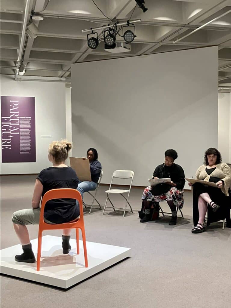 Artists drawing on boards surround a live figure model who poses on a platform in an orange chair.