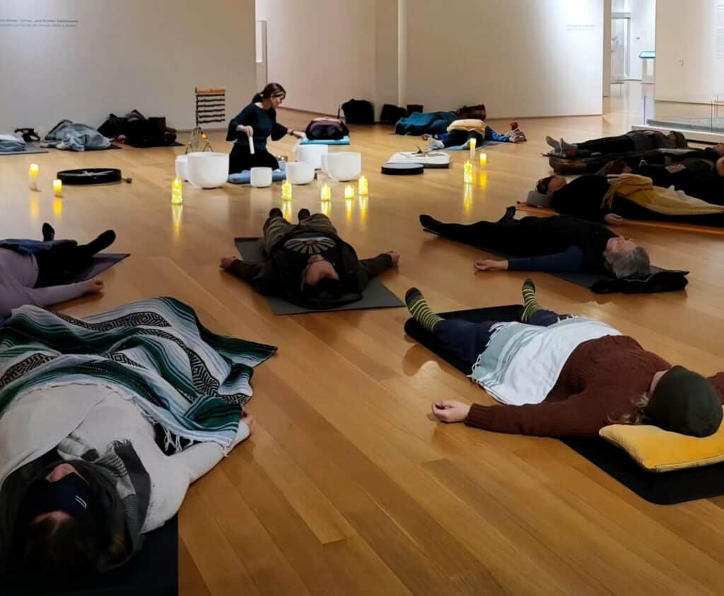 A group of people lay on the floor in a circle in the galleries, on mats and wrapped in blankets with their eyes closed while a facilitator plays vibrational instruments like crystal singing bowls.