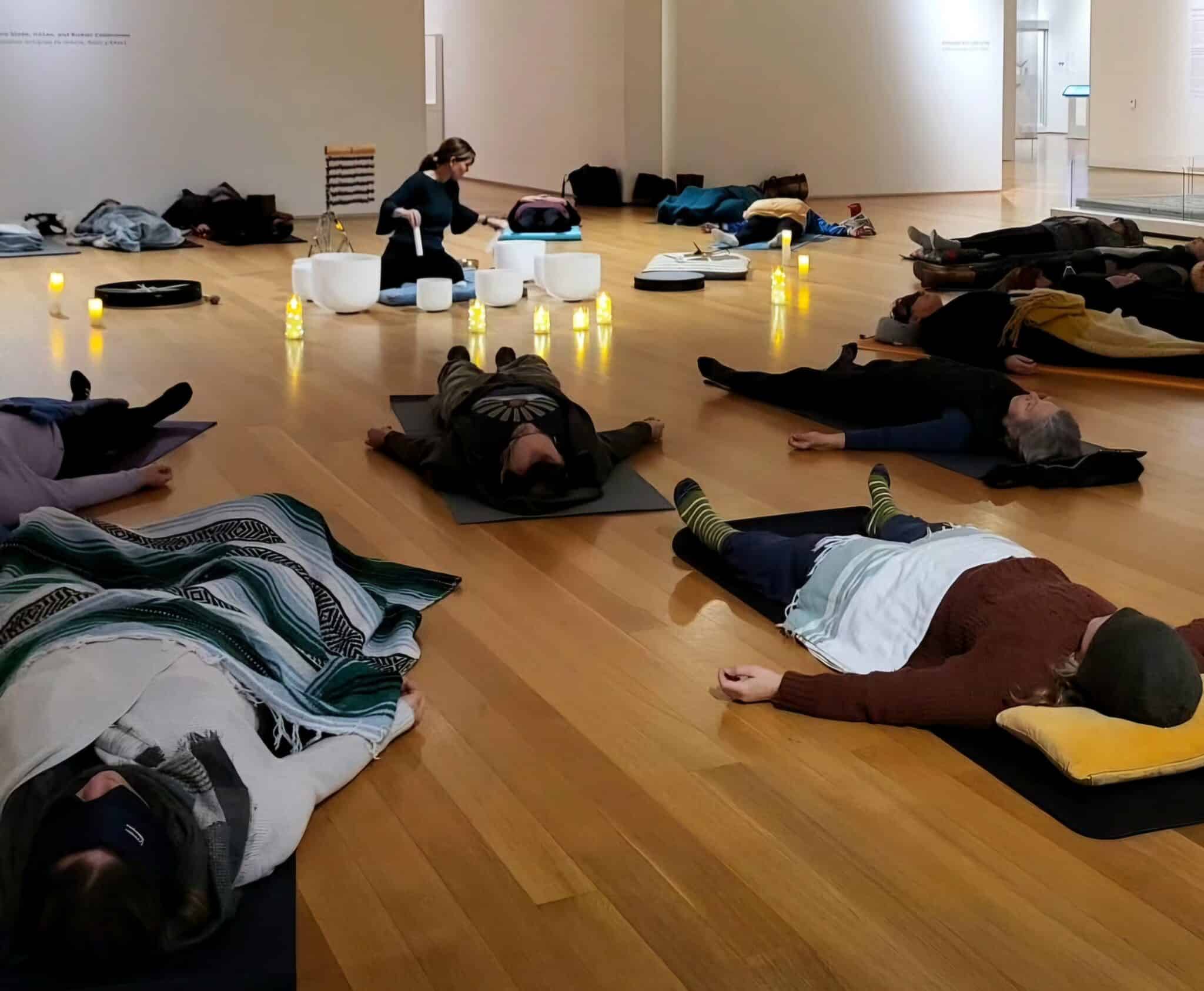 A group of people lay on the floor in a circle in the galleries, on mats and wrapped in blankets with their eyes closed while a facilitator plays vibrational instruments like crystal singing bowls.
