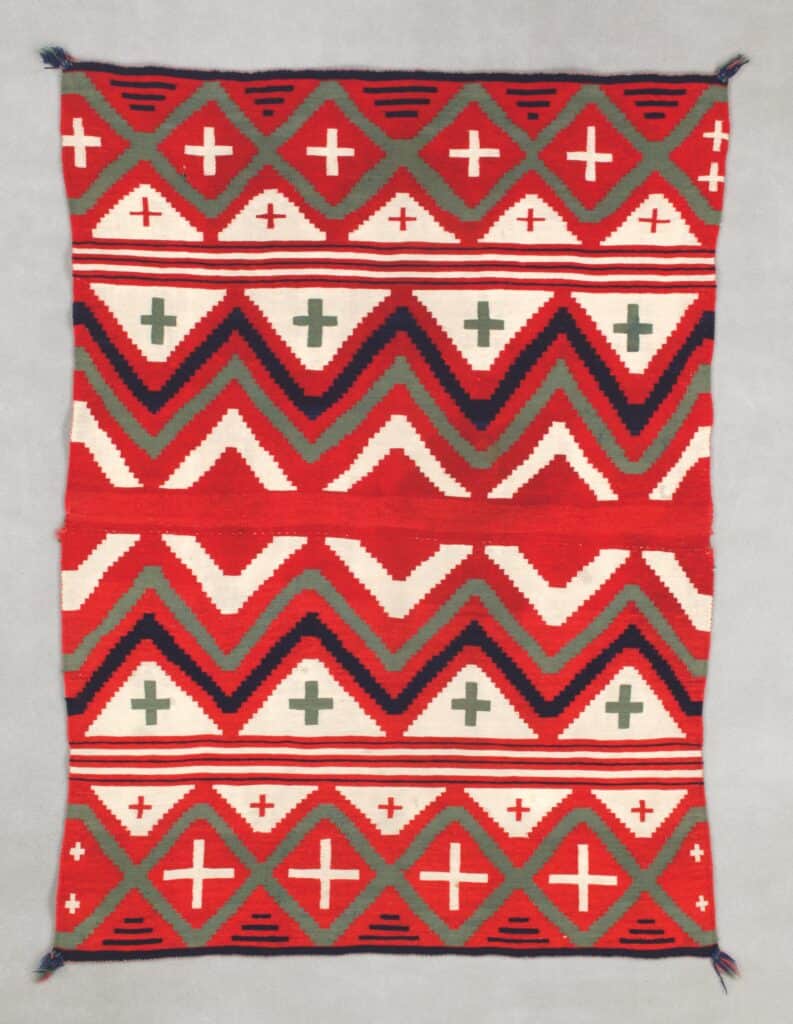 A rectangular blanket with a geometric pattern containing orange, white, and black zigzags and crosses.