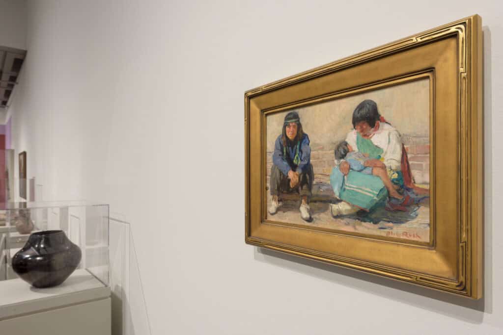 An oil painting of two figures sitting on a curb hangs in a gold frame next to a piece of blackware pottery encased in a glass display.