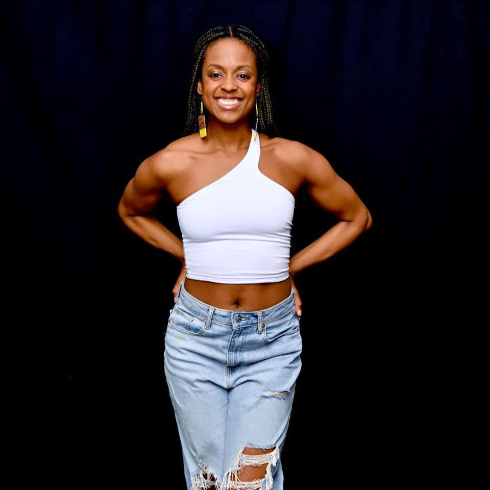 Photograph of Jordan Farrell dressed in jeans and a white tank top smiling and standing against a black backdrop.