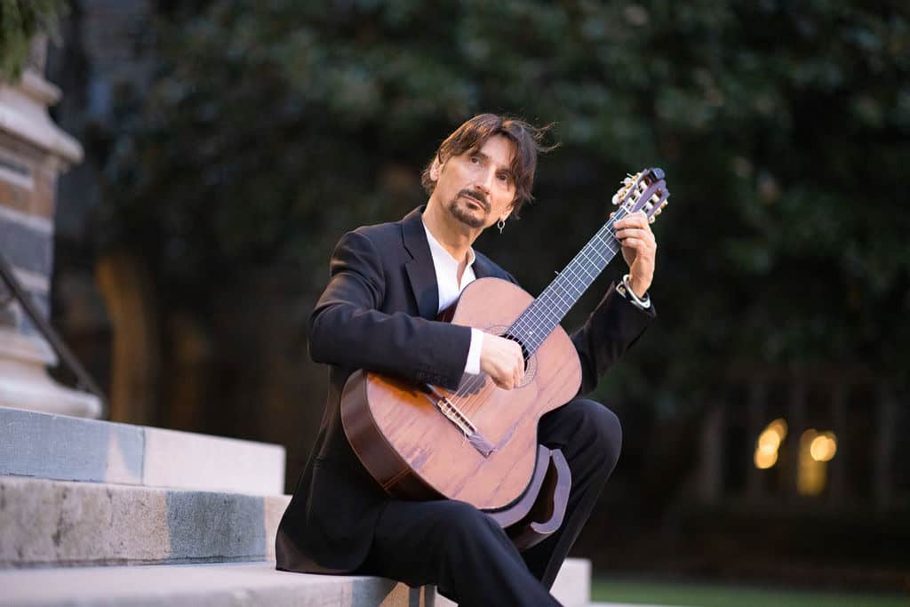 A man seated on outdoor granite steps strums a guitar and looks up and to the left.