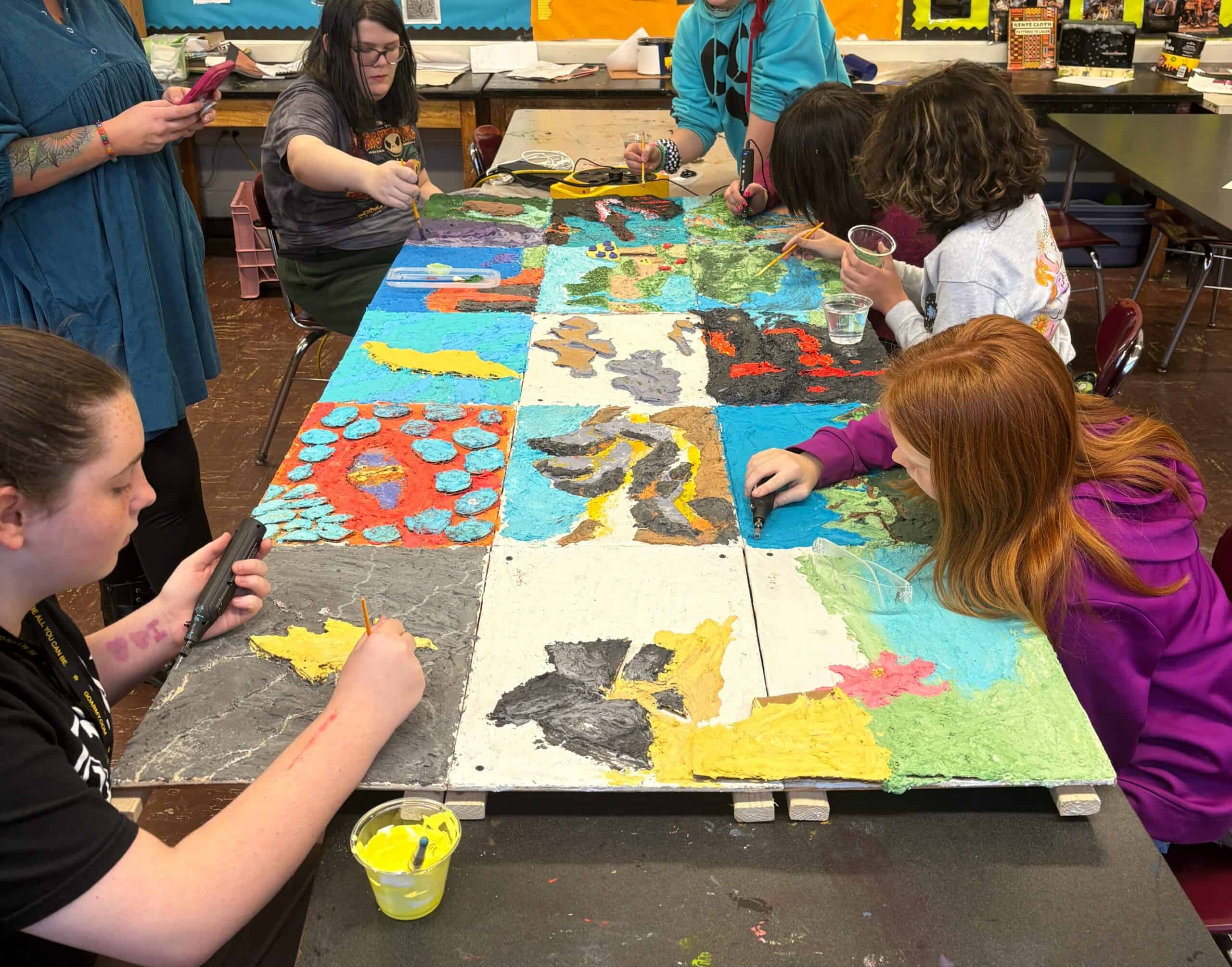 In a classroom setting, youths paint an artwork comprised of 15 individual rectangular boards nailed together.