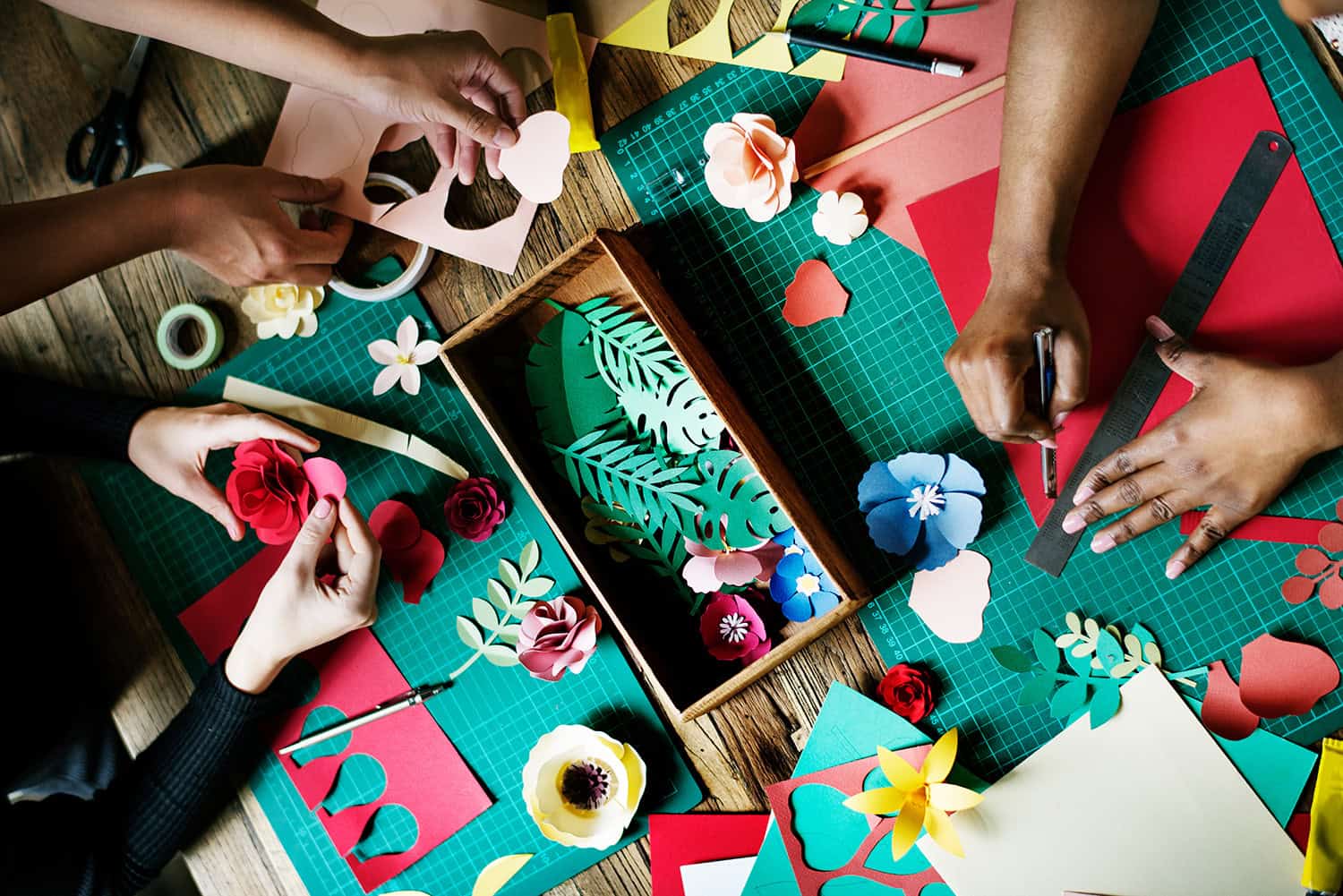 An overhead image of three sets of hands creating paper flowers over a table of art supplies.