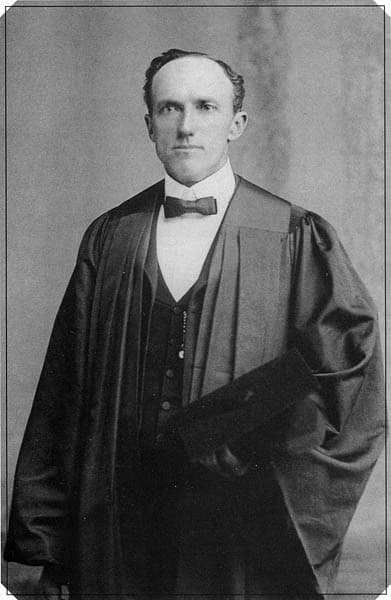 A black-and-white photograph of a man with short hair posing in a graduation gown. Underneath the gown, he wears a vest, dress shirt, and bowtie. He holds a graduation cap with his left hand. 