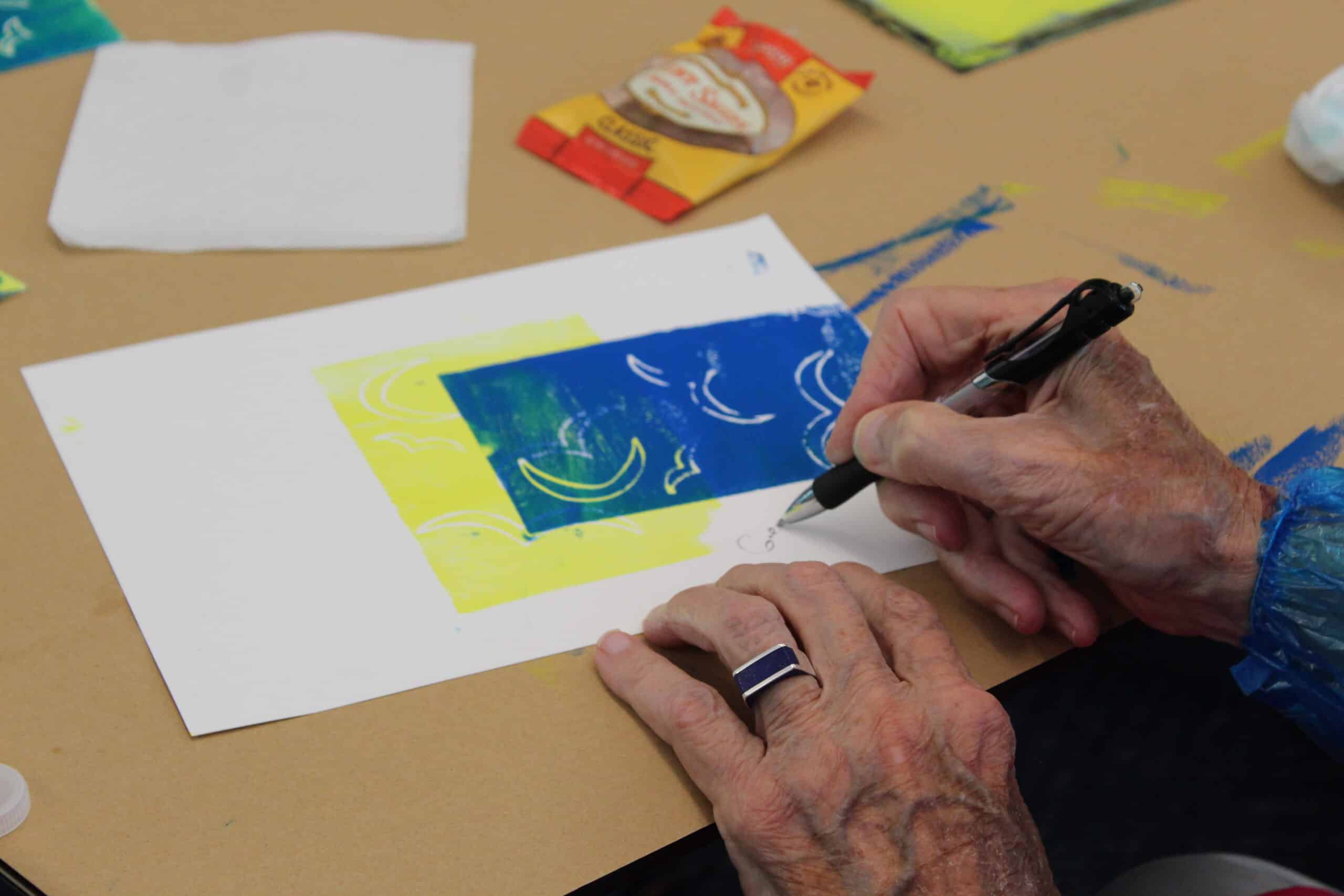 Close-up of hands that show wrinkles and veins. Right hand writes with a pen on paper printed with a moon and birds.